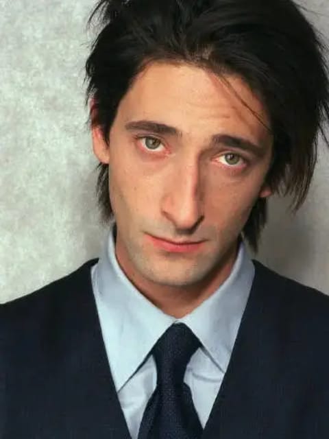 Retrato de Adrien Brody com cabelo preto com textura bagunçada. Ele usa terno preto combinado com camisa azul clara e gravata preta. Modelo para troca de rosto de imagem.