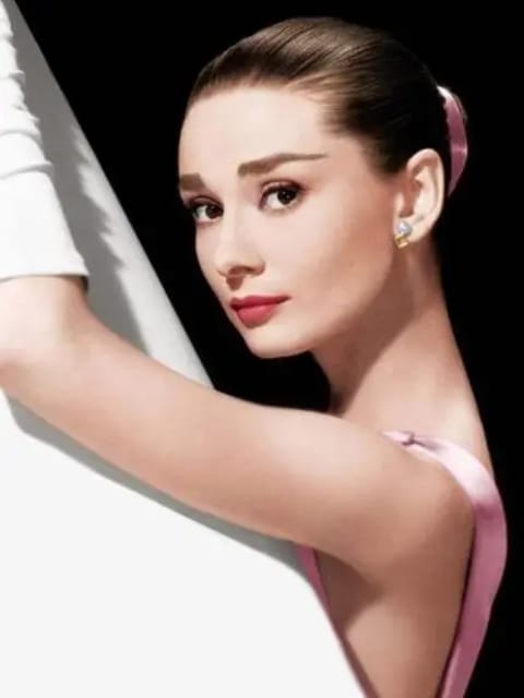 Retrato de Audrey Hepburn com penteado elegante puxado para trás adornado com acessório de cabelo rosa, brincos de pérola e vestido rosa suave sem mangas. Ela se apoia em uma estrutura geométrica branca com fundo preto. Modelo para troca de rosto de imagem.