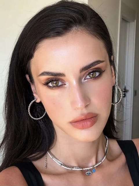 Selfie de uma mulher com cabelo preto liso, brincos de argola prateada e colar, posando em uma cena interna. Modelo para troca de rosto de imagem.