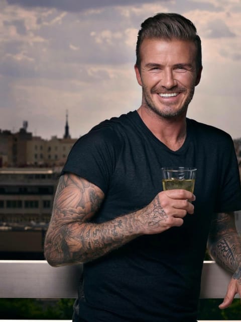 Foto média de David Beckham em pé em uma varanda com a paisagem urbana de Barcelona como pano de fundo, ele tem um corte inferior texturizado elegante com um lado desbotado e cabelo penteado para trás, tatuagens de manga comprida em ambos os braços, usa uma camiseta preta com decote em V, segura um copo com uma bebida amarela e sorri amplamente para a câmera.