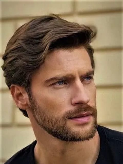 Um retrato em close de um homem com cabelos castanhos grossos e penteados, com ondas suaves e sutis reflexos castanhos claros. Ele usa uma blusa preta de gola redonda, posicionada em frente a uma parede de azulejos bege claro, olhando fora da câmera com uma expressão calma. Modelo para troca de rosto de imagem.