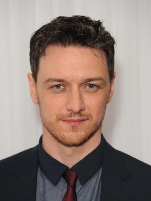 Retrato de James McAvoy com cabelo escuro curto e ondulado com mechas ruivas e grisalhas. Ele usa um blazer azul marinho escuro sobre uma camisa xadrez azul clara, uma gravata vermelha escura e uma camiseta de colarinho escuro, contra um fundo texturizado branco liso.