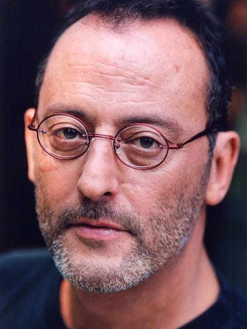 Close de Jean Reno apresentando seu cabelo curto e escuro com a linha do cabelo recuando, óculos redondos de aro metálico com armação avermelhada, barba e bigode grisalhos. Ele usa um top azul escuro com gola redonda e olha calmamente para a câmera.