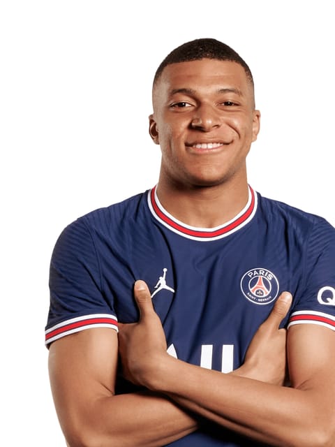 Retrato de Kylian Mbappe com um corte curto e vestindo a camisa azul do Paris Saint-Germain. ele está com os braços cruzados e sorri para a câmera contra um fundo branco e liso.