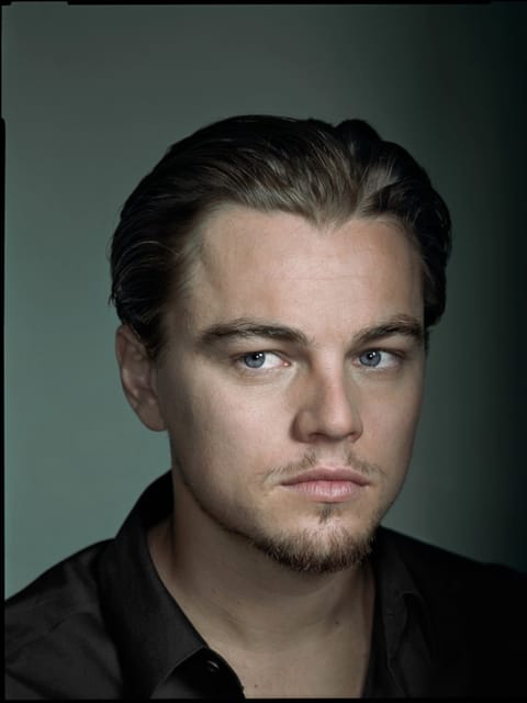 Retrato de Leonardo Dicaprio com costas lisas e camisa preta, posando em estúdio mal iluminado com fundo verde, olhando fora da câmera com expressão calma. Modelo para troca de rosto de imagem.
