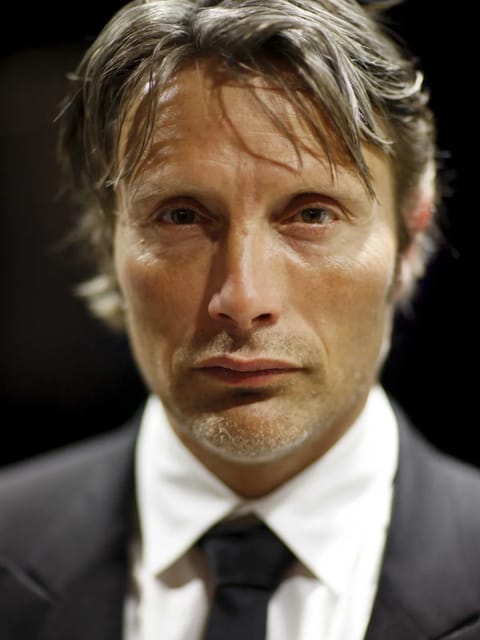 Retrato de Mads Mikkelsen com ondas texturizadas bagunçadas, em terno preto com gravata preta, olhando para a câmera com expressão calma.
