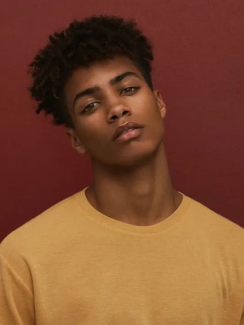 Retrato de homem com cabelo afro preto e expressão calma, vestindo blusa amarela com gola careca e cabeça inclinada para o lado. Modelo para troca de rosto de imagem.