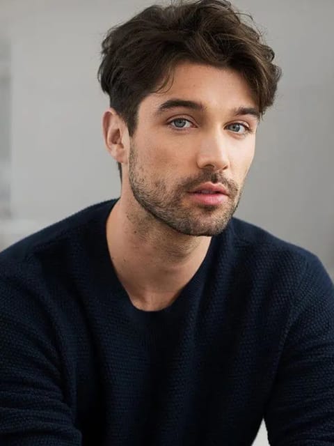 Retrato de um homem com cabelos castanhos escuros grossos e ondulados, com aparência de textura bagunçada, vestindo um suéter azul marinho, sentado em uma cena interna com expressão calma. Modelo para troca de rosto de imagem.