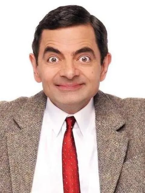 Retrato do Sr. Bean com cabelo penteado para o lado e expressão exagerada, vestindo um blazer cáqui sobre uma camisa de colarinho branco e uma gravata vermelha. Seus braços se abrem, contra um fundo branco e liso.