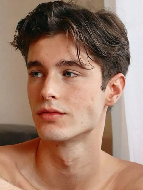 Retrato de um homem sem camisa, com cabelos castanhos ondulados e grossos, sentado em uma cena interna, olhando fora da câmera com uma expressão calma. Modelo para troca de rosto de imagem.
