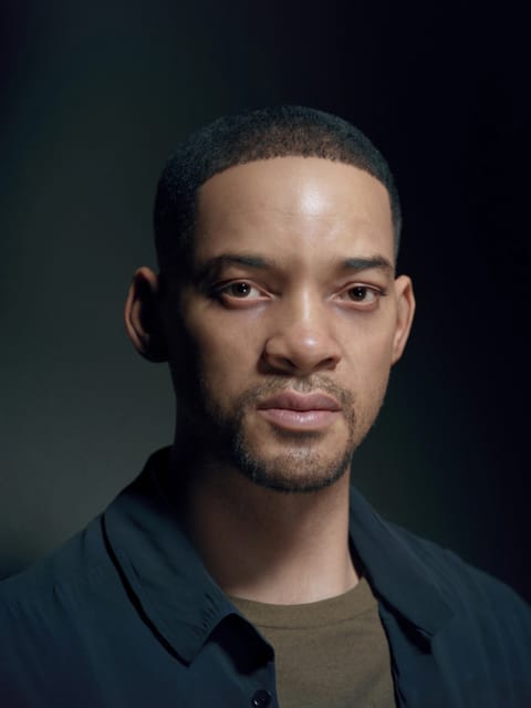 Retrato de Will Smith com seu cabelo curto e escuro e barba por fazer, ele veste uma camisa de colarinho desabotoado azul marinho sobre uma camiseta bege com gola redonda e olha diretamente para a câmera contra um fundo gradiente cinza escuro suave. Modelo para troca de rosto de imagem.