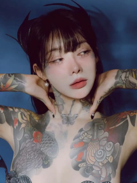 Retrato de uma mulher de cabelos pretos com franja romba e tatuagem floral de inspiração japonesa no peito e ombro. Ela está posando na água com as duas mãos na nuca. Modelo para troca de rosto de imagem.