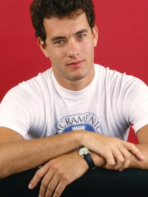 Retrato de estúdio do jovem Tom Hanks com cabelo curto, escuro e cacheado. Ele veste uma camiseta branca com gola redonda e estampa gráfica azul, descansa os braços cruzados sobre uma superfície, tem um relógio de pulso no pulso esquerdo e olha para a câmera contra um fundo vermelho sólido e brilhante. Modelo para troca de rosto de imagem.
