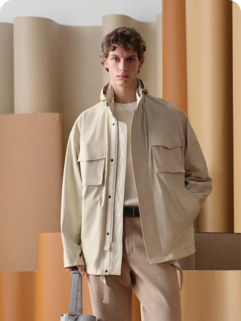 Un hombre con una chaqueta beige y pantalones a juego, sosteniendo un bolso gris, en un ambiente interior moderno, modelo para el intercambio de ropa de IA.