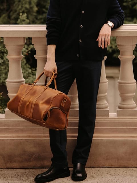 Un hombre con un cárdigan y pantalones negros, sosteniendo un bolso marrón, en un jardín, modelo para el intercambio de ropa de IA.
