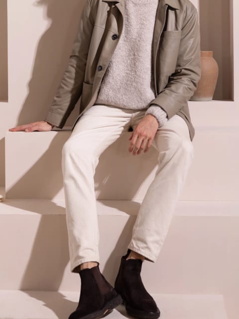 Un hombre con una chaqueta gris, un suéter gris claro y pantalones blancos, sentado en un espacio minimalista, modelo para el intercambio de ropa de IA.