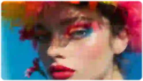 Foto buram seorang wanita yang menampilkan perona mata biru dan oranye, lipstik merah, dan hiasan kepala berbulu warna-warni.