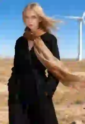 Portrait of a blonde woman in a long black knitted trench coat paired with a flowing brown-beige scarf, standing in a wind farm field. Her hair flying in the wind. A template for image to image tool.