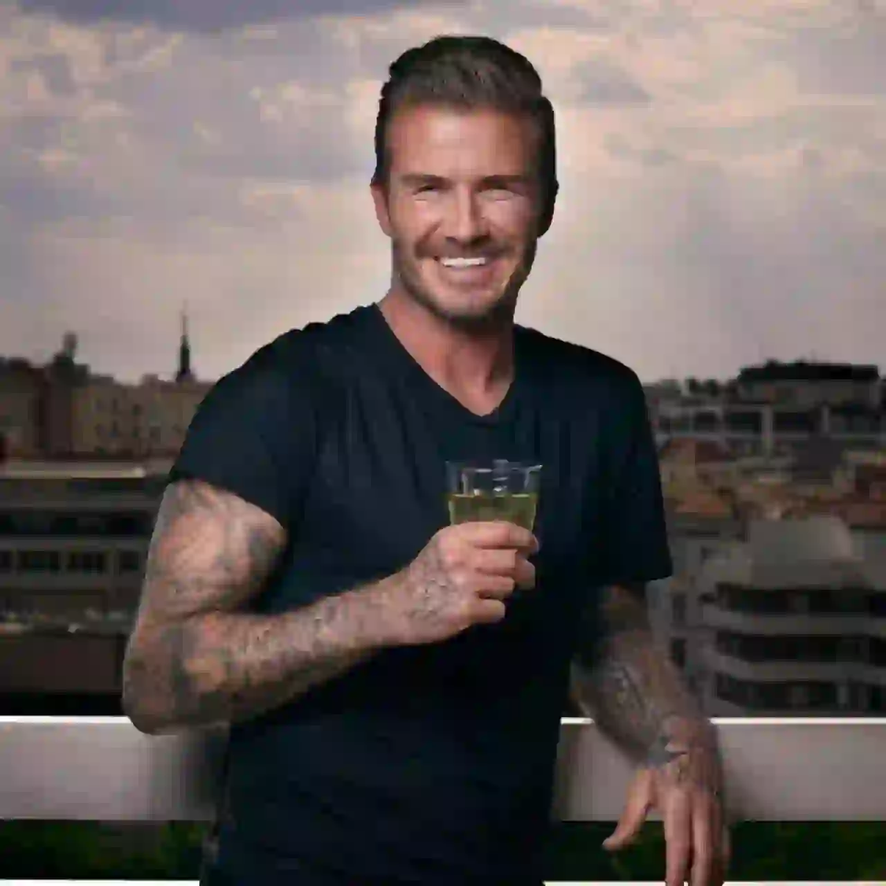 Medelstor bild av David Beckham som står på en balkong med Barcelonas stadsbild i bakgrunden, han har en stilig texturerad underskärning med en blek sida och bakåtsvept hår, tatueringar med helärm på båda armarna, bär en svart t-shirt med V-ringad, håller ett glas med en gul dryck och flinar brett mot kameran.