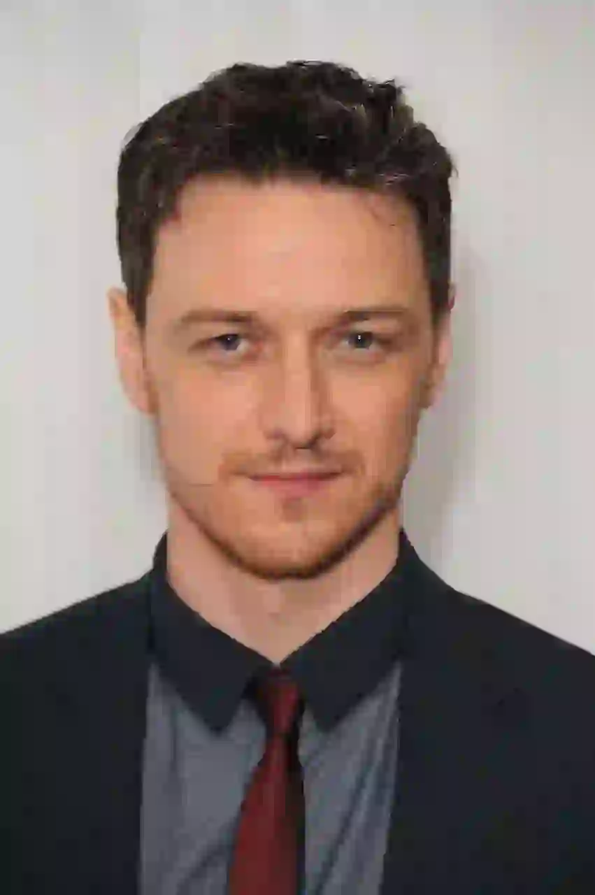 Porträtt av James McAvoy med kort vågigt mörkt hår streckat med grå, röd ingefära stubb. Han bär en mörk marinblå kavaj över en ljusblå rutig skjorta, en mörkröd slips och en undertröja med mörk krage, mot en vanlig vit texturerad bakgrund.