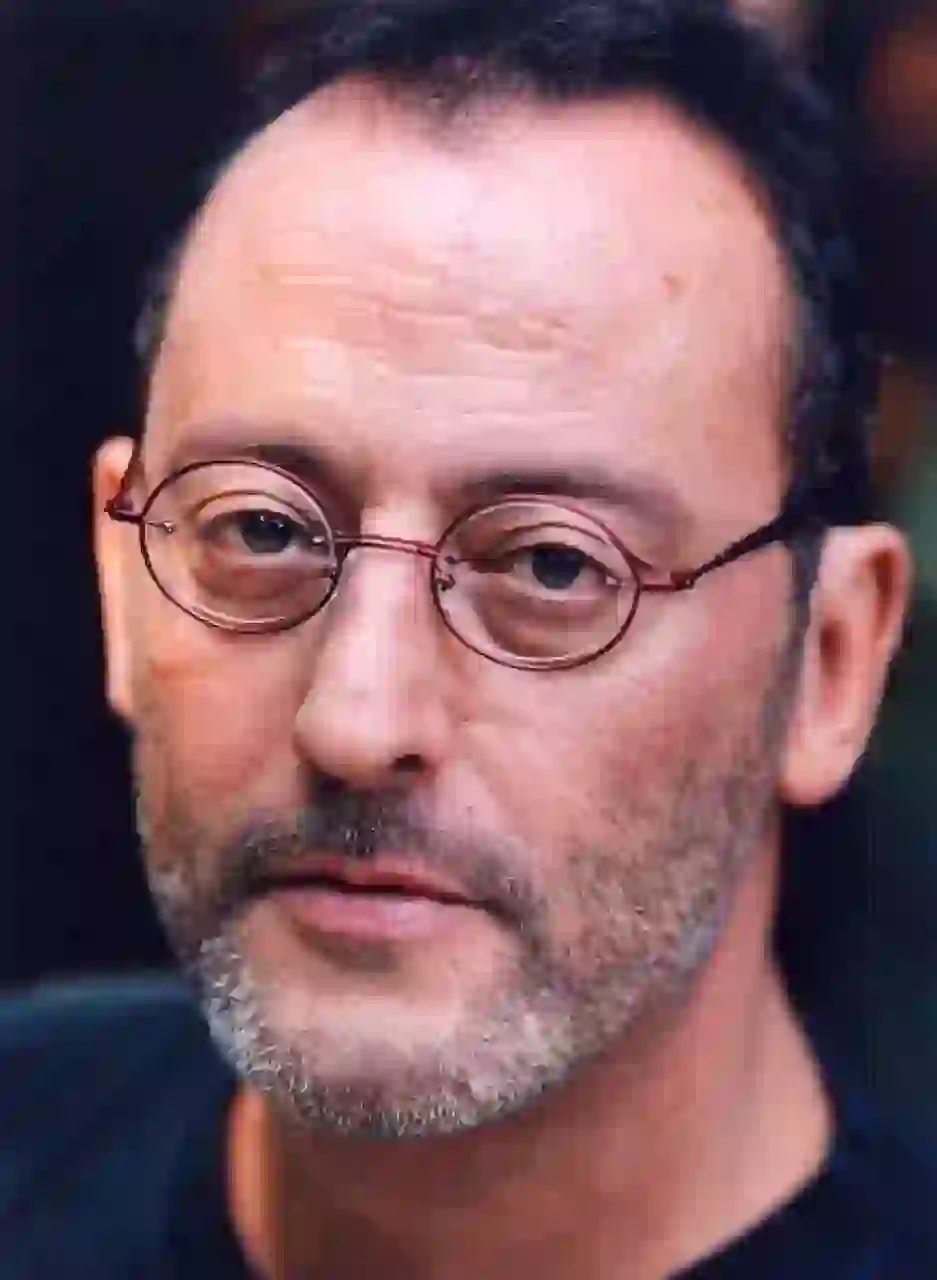 Närbild av Jean Reno med sitt korta mörka hår med ett vikande hårfäste, runda glasögon med rödbågade glasögon med en rödaktig båge, ett skägg med salt och peppar stubb och mustasch. Han bär en mörkblå topp med rund hals och stirrar lugnt mot kameran.