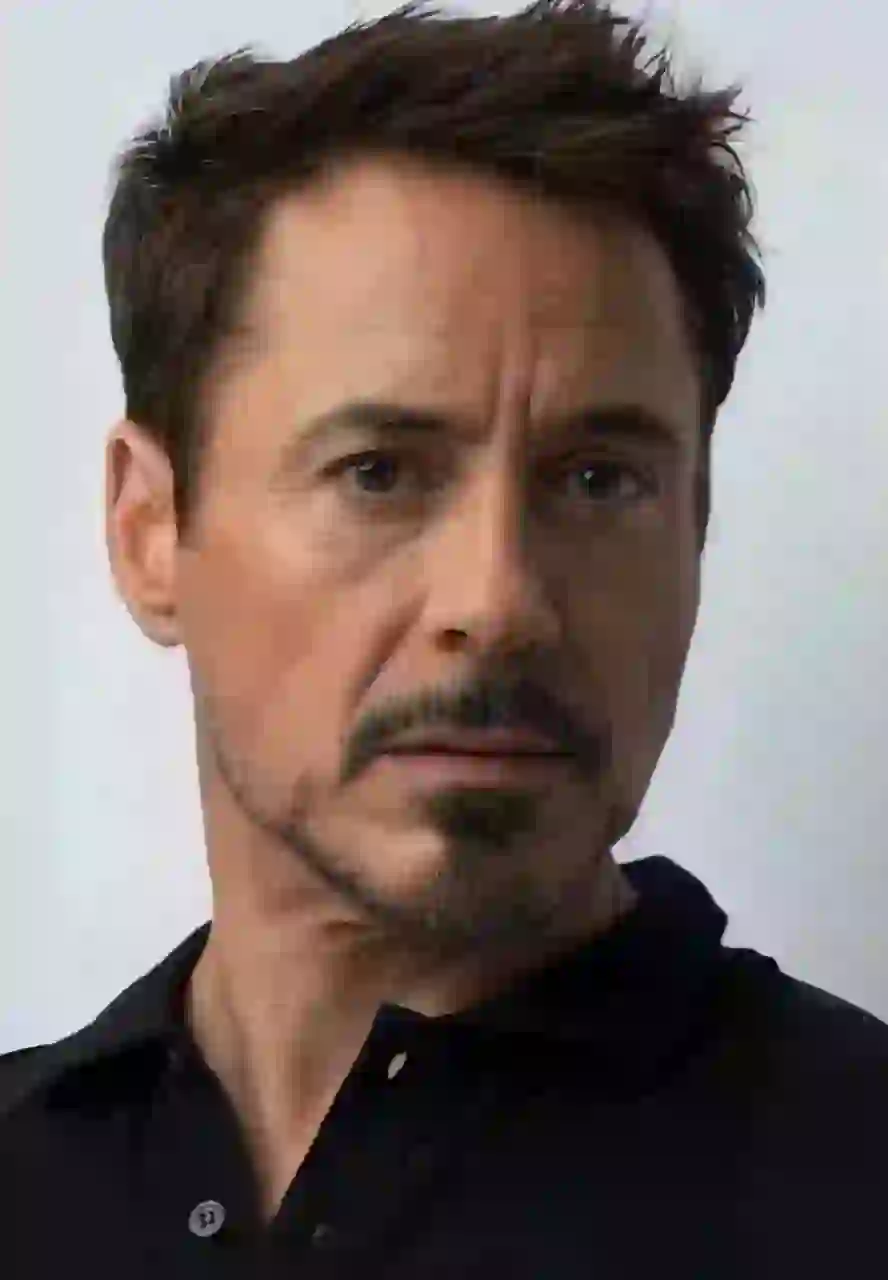 Närbild av Robert Downey Jr. med kort rörigt mörkt hår streckat med grått, ett trimmat bockskägg och mustasch med salt- och peppartoner. Han bär en skjorta med svart krage med en delvis uppknäppt knappslå och stirrar direkt på kameran mot en vanlig vit bakgrund. Mall för bildbyte av ansikte.