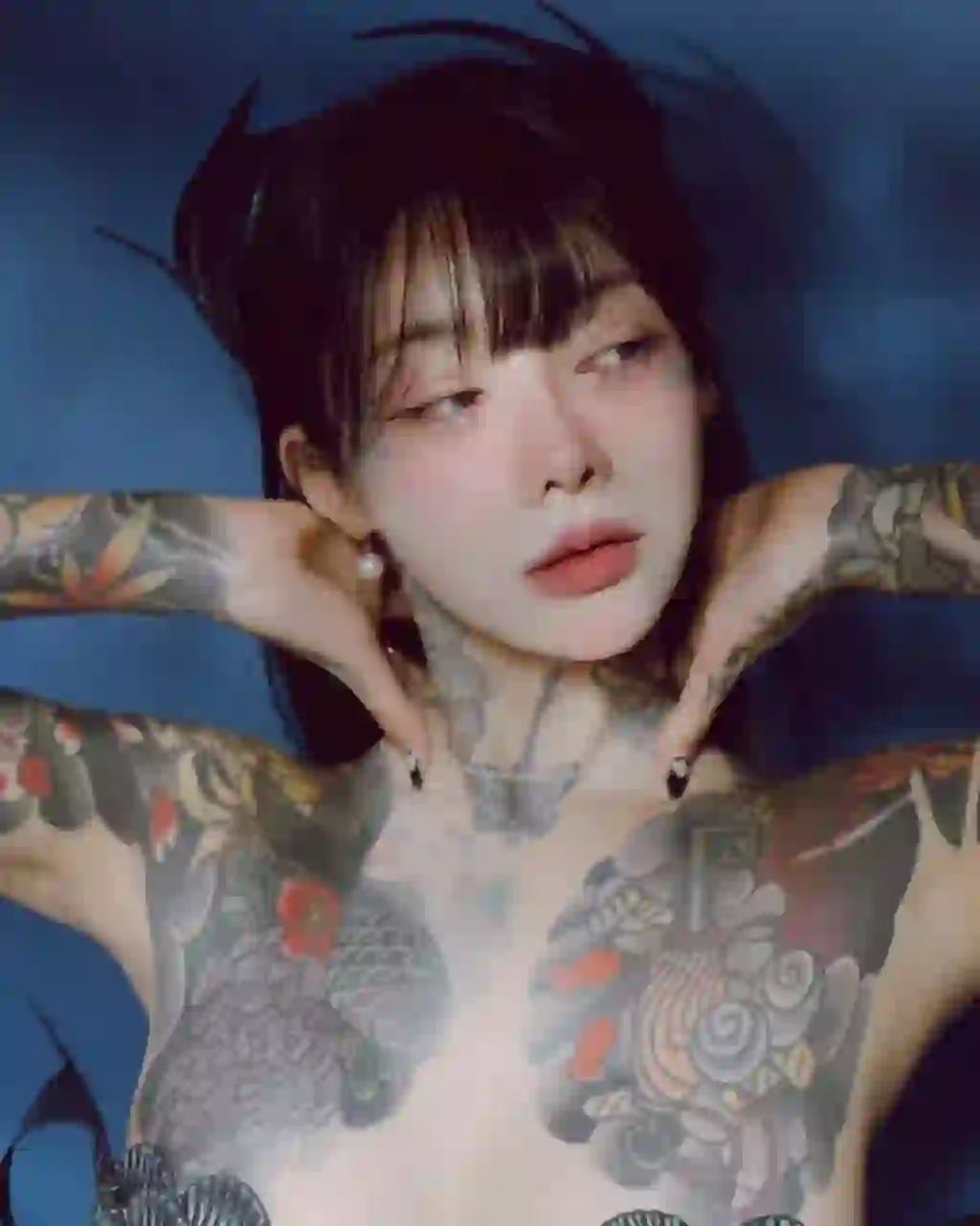 Portrait d'une femme aux cheveux noirs avec une frange émoussée et un tatouage floral d'inspiration japonaise sur la poitrine et l'épaule. Elle pose dans l'eau, les deux mains sur la nuque. Modèle pour l'échange de visage d'image.