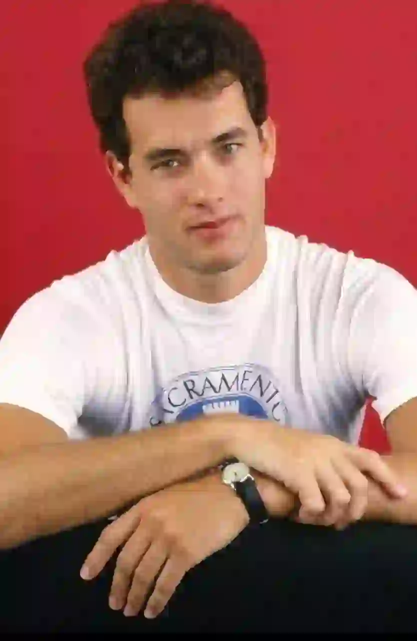 Studioporträtt av unge Tom Hanks med kort tjockt mörkt lockigt hår. Han bär en vit t-shirt med rund hals och blått grafiskt tryck, vilar armarna i kors på en yta, har ett armbandsur på sin vänstra handled och stirrar på kameran mot en fast ljusröd bakgrund. Mall för bildbyte av ansikte.