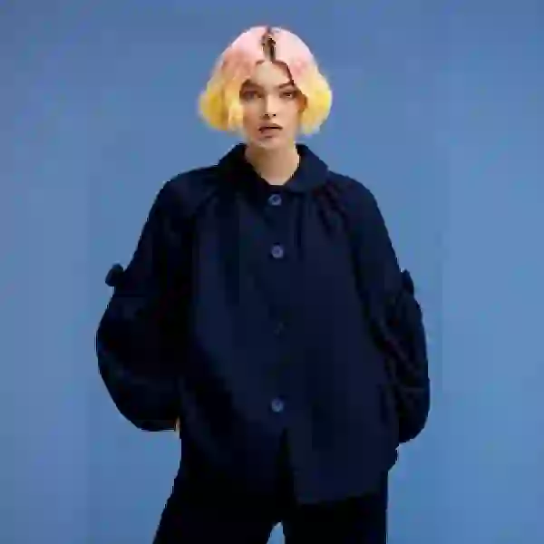 Bức ảnh hoán đổi khuôn mặt của một người phụ nữ với mái tóc hồng vàng, mặc áo sơ mi đen và quần đen, tạo dáng trước ống kính.