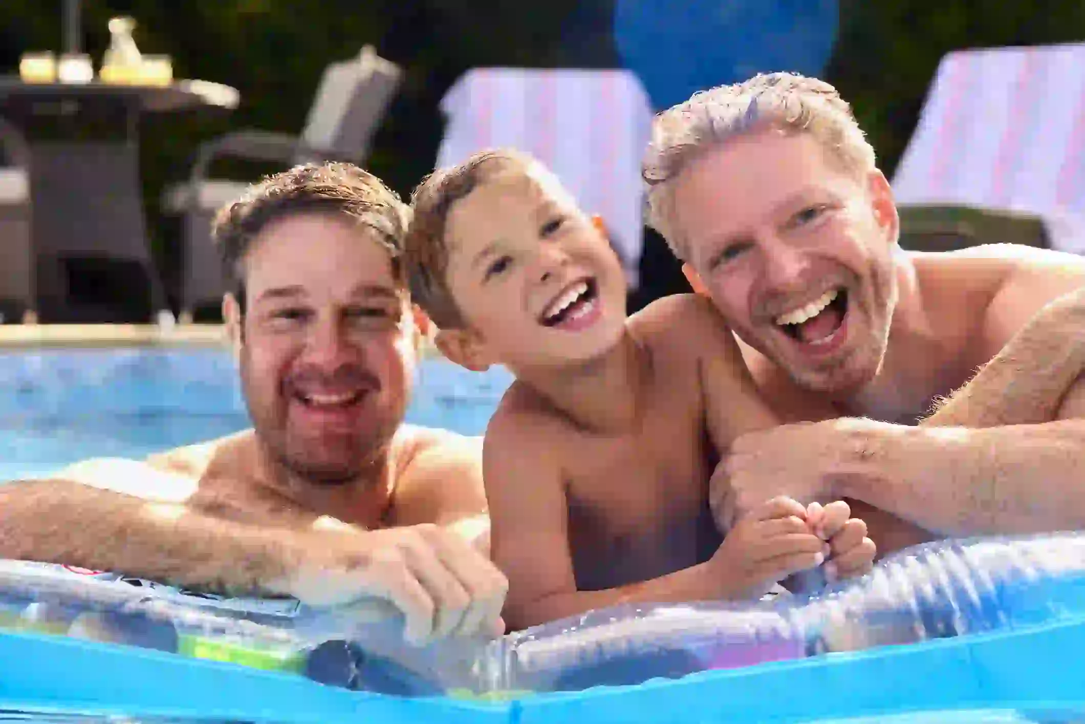 This is a screenshot of the gay dads and their son after the multiple face swap video. The three of them are still playing by the pool, laughing. The gay dads' faces have been swapped with Cam and Mitch from Modern Family, and the son's face has been swapped with Luke Dunphy.