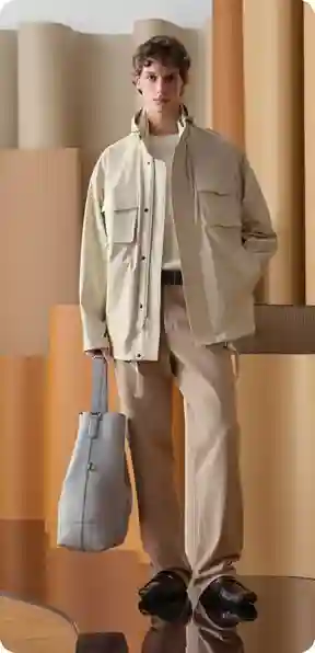 En mand i en beige jakke og matchende bukser, holder en grå taske, i et moderne interiør, model for AI-tøjbytte.