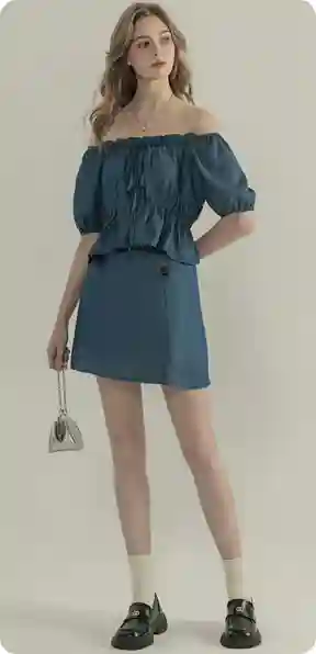 En kvinde i en blå top med skulderåbning og matchende nederdel med en lille grå taske, model for AI-tøjbytte.